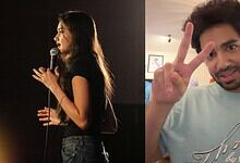 Young woman performing stand-up comedy on stage with microphone, man making peace sign in background.