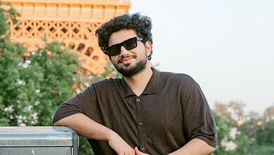 Young man with sunglasses and beard smiling outdoors in front of a monument and greenery.