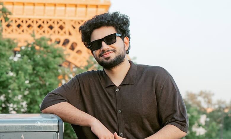 Young man with sunglasses and beard smiling outdoors in front of a monument and greenery.