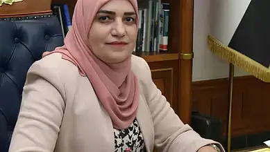 Iraqi woman wearing a pink hijab and beige blazer in an office setting.