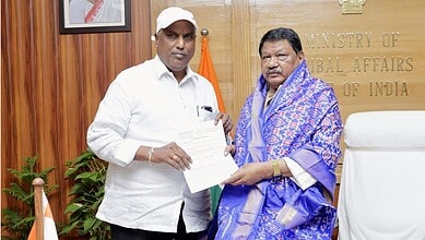 Two men in an office, one presenting a document to the other, with a Ministry of Tribal Affairs sign in a.