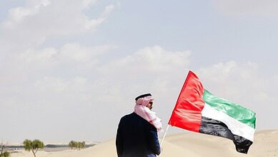 Person holding UAE flag in sandy desert with blue sky and clouds.