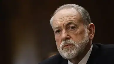 Mike Huckabee speaking at a public event, serious expression, grey beard, dark background.