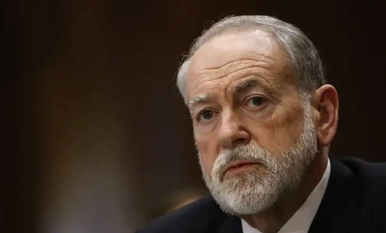 Mike Huckabee speaking at a public event, serious expression, grey beard, dark background.