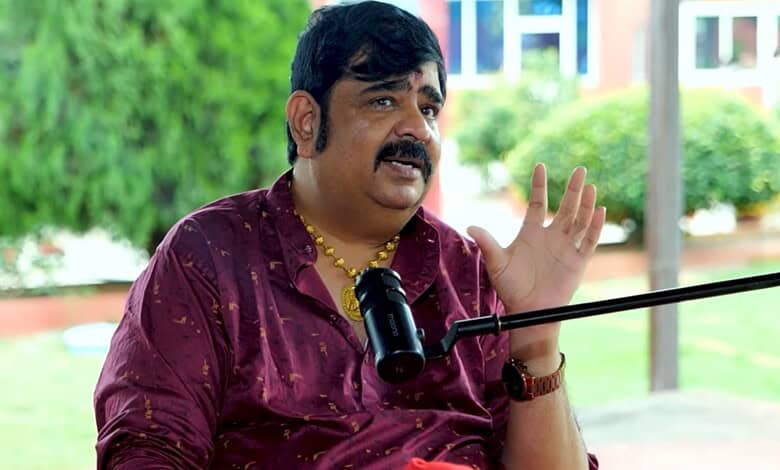 Astrologer Venu Swamy speaking outdoors with a microphone, gesturing with his hand.