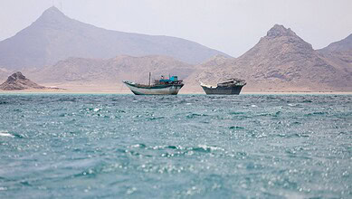 Two boats in rough waters near mountainous coastline, Yemen, during daytime.
