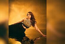 A woman in a black dress with a floral top poses on a reflective surface against a golden background.