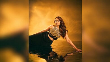 A woman in a black dress with a floral top poses on a reflective surface against a golden background.
