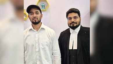 Two men, one in a white shirt and the other in a black robe, standing indoors with a pink wall background.