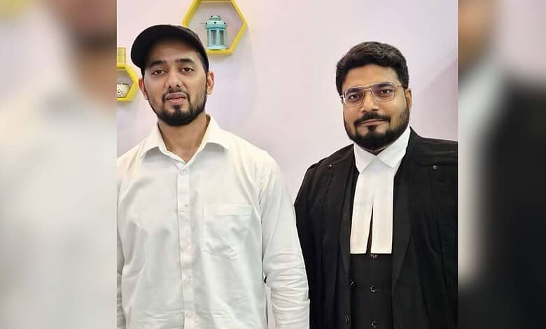 Two men, one in a white shirt and the other in a black robe, standing indoors with a pink wall background.