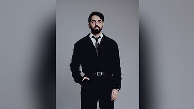 Ayushmann Khurrana wearing a black suit and tie, posing against a grey background.