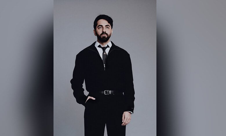 Ayushmann Khurrana wearing a black suit and tie, posing against a grey background.