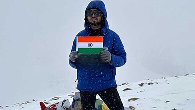 Man in blue jacket holding Indian flag on snowy mountain peak during daytime.
