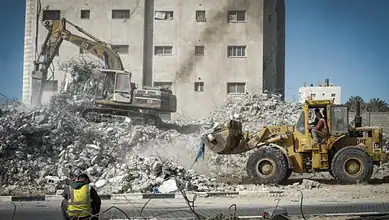 Bulldozers demolish rubble in Gaza, with workers observing the destruction.