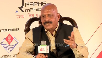 Man speaking at a conference with a microphone, wearing a beige shirt and black vest.