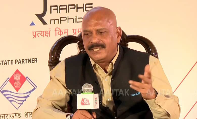 Man speaking at a conference with a microphone, wearing a beige shirt and black vest.