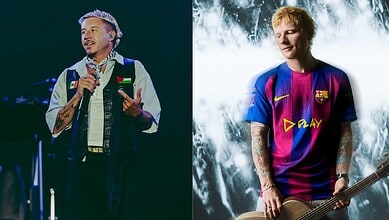 Rapper Macklemore performing on stage with a microphone, wearing a denim vest and patterned shirt.