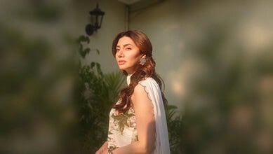 Mahira Khan with long wavy hair, wearing a white dress, standing outdoors in sunlight.