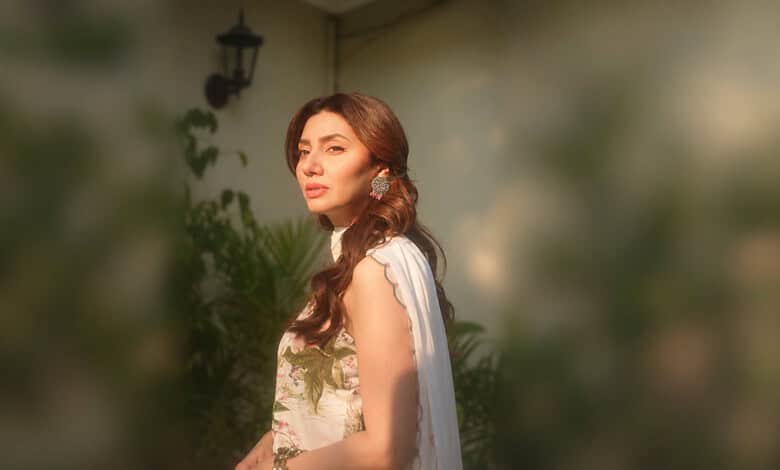 Mahira Khan with long wavy hair, wearing a white dress, standing outdoors in sunlight.