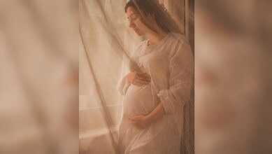 Pregnant woman standing by window, gently holding her belly, soft natural light illuminating her.