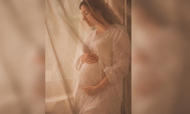 Pregnant woman standing by window, gently holding her belly, soft natural light illuminating her.