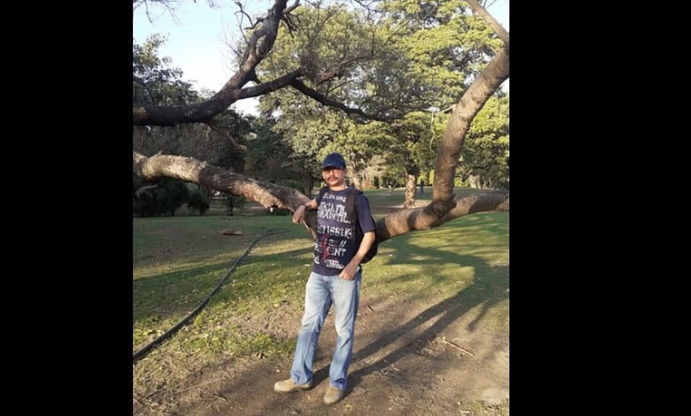 Man in casual clothes standing under a large, sprawling tree in a park on a sunny day.