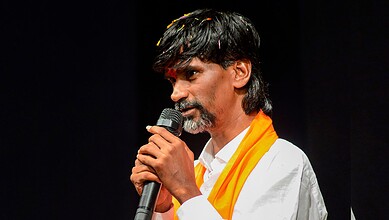 Man with black hair and beard speaking into a microphone, wearing a white shirt and orange scarf.