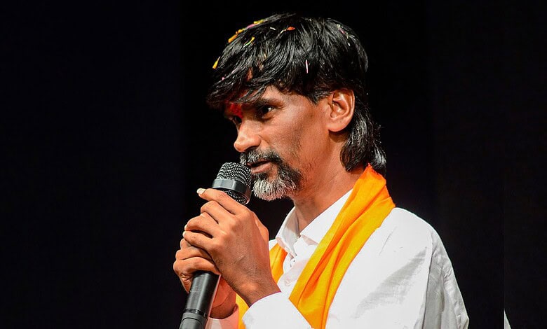 Man with black hair and beard speaking into a microphone, wearing a white shirt and orange scarf.