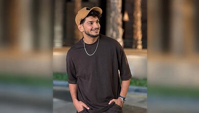 Young man in black t-shirt and beige cap smiling outdoors at night.