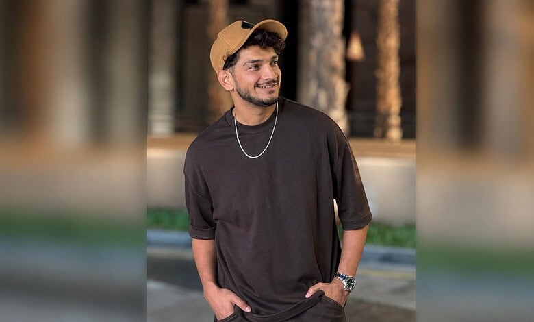 Young man in black t-shirt and beige cap smiling outdoors at night.