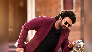 Prabhas in a maroon blazer and sunglasses, smiling and looking at his phone outdoors.