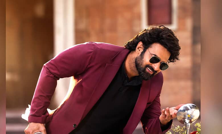 Prabhas in a maroon blazer and sunglasses, smiling and looking at his phone outdoors.