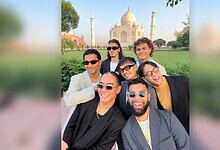 Group of diverse friends smiling in front of the Taj Mahal during daytime.