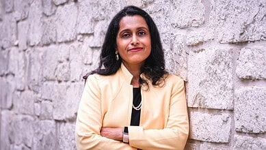 Indian woman in cream blazer standing against stone wall, smiling confidently.