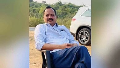 Man sitting outdoors near a white car, wearing a light blue shirt and dark trousers.