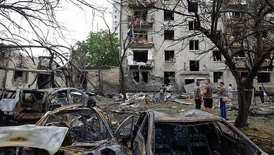 Damaged buildings and burnt-out cars in a war-torn area with people inspecting the scene.