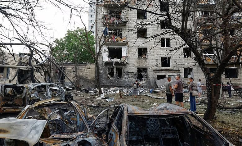 Damaged buildings and burnt-out cars in a war-torn area with people inspecting the scene.
