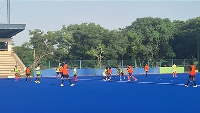Young athletes training on a blue outdoor hockey field with coaches and lush green trees in the backdrop.