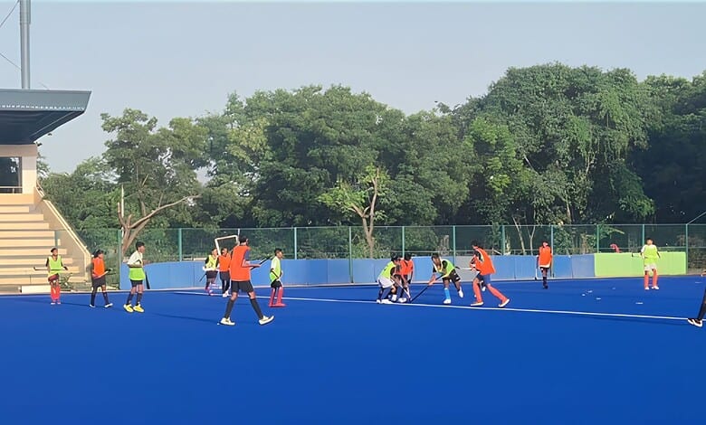 Young athletes training on a blue outdoor hockey field with coaches and lush green trees in the backdrop.