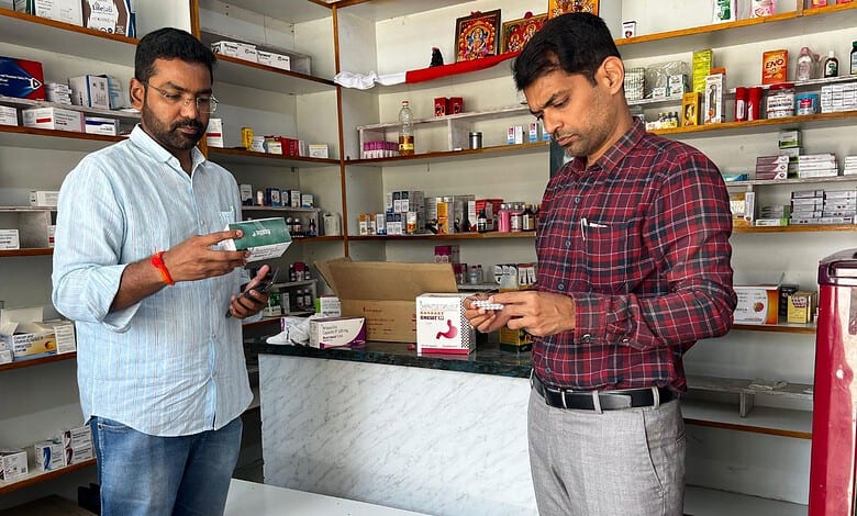 Two men inspecting medicines in a pharmacy, shelves filled with drugs in the background.