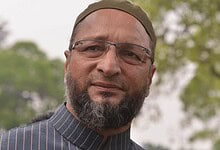 Owaisi speaking outdoors with trees in the background, wearing glasses and a cap.