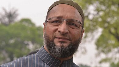 Owaisi speaking outdoors with trees in the background, wearing glasses and a cap.