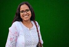 Smiling woman with glasses and a white floral kurta against a green background.