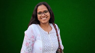 Smiling woman with glasses and a white floral kurta against a green background.