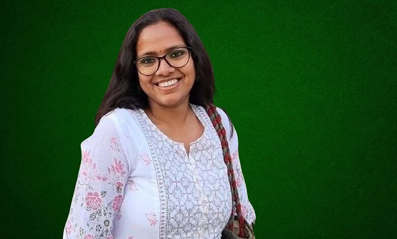 Smiling woman with glasses and a white floral kurta against a green background.