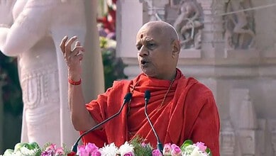 Govind Dev Giri speaking at a religious event with a temple backdrop.