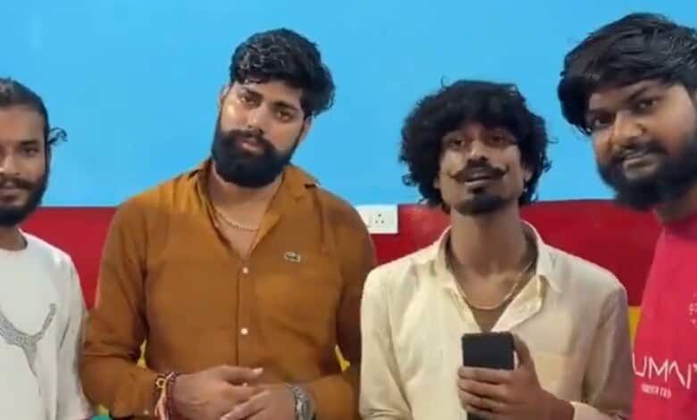 Group of four young men standing against a blue and red wall, one holding a smartphone.