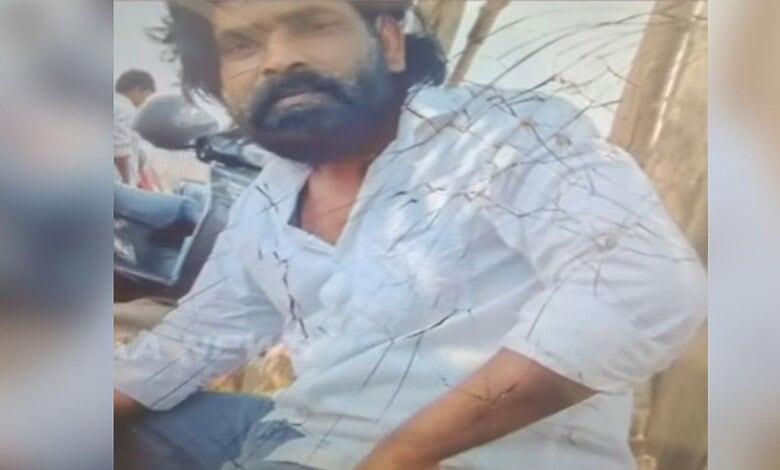 Man with a beard and long hair sitting outdoors, wearing a white shirt with visible cracks.