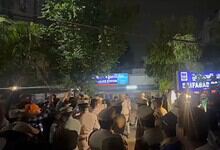 Crowd gathered outside a police station at night with illuminated signs and trees in the background.