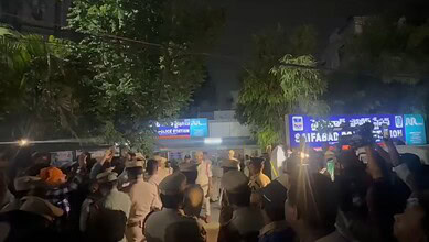 Crowd gathered outside a police station at night with illuminated signs and trees in the background.
